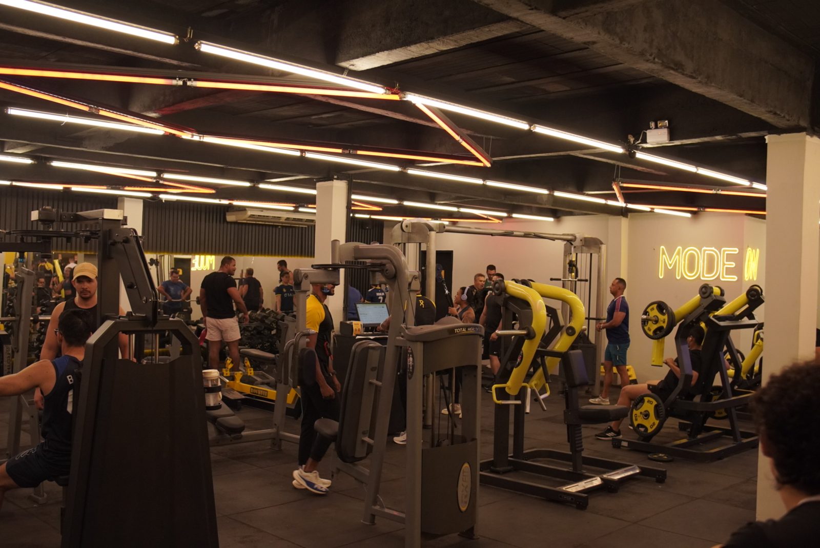 Salão de musculação lotado com máquinas amarelas e letreiro neon Mode On na unidade Abreu e Lima