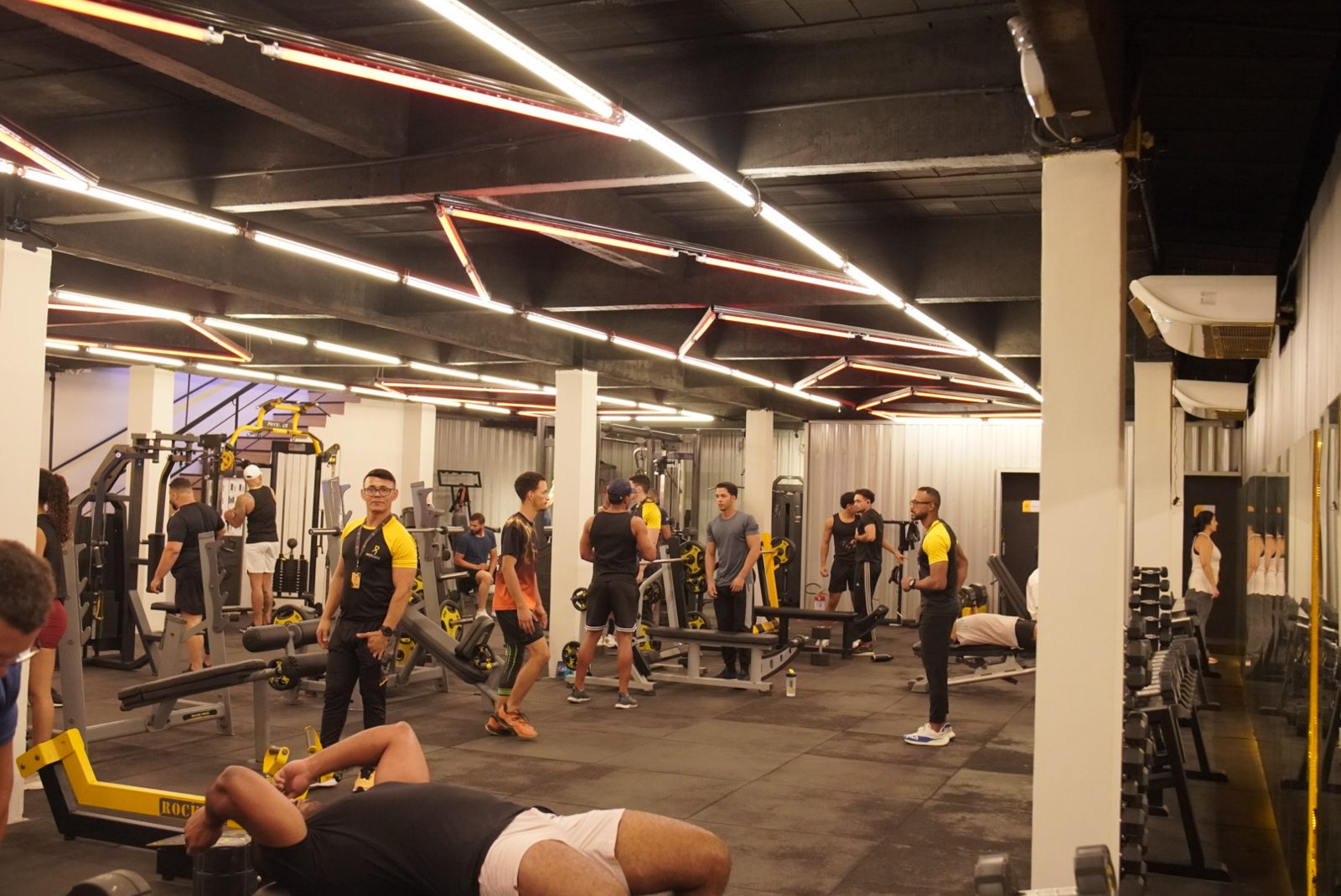 Amplo salão de musculação com bancos, halteres e alunos treinando na unidade Abreu e Lima